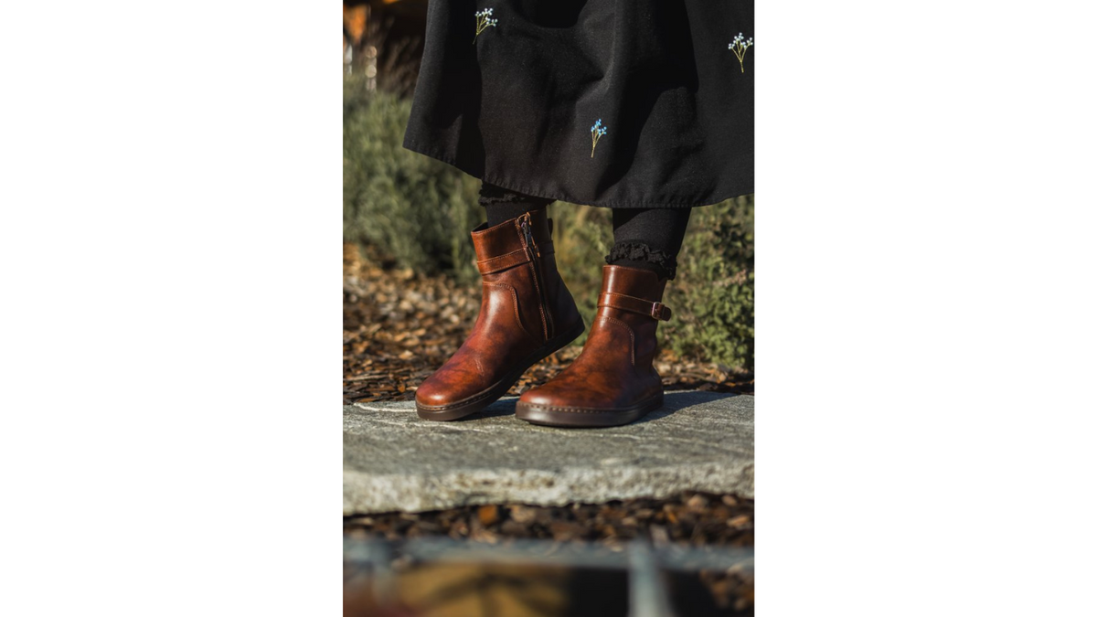 Barfußstiefel am Fuß getragen mit seitlichem Reißverschluss und Riemen, breiter Zehenbox und flacher Sohle, geschlossener Fersenbereich, schräge Vorderansicht