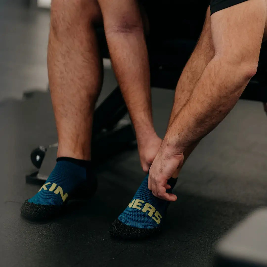 Person zieht schwarze Skinners Socken mit gelbem Logo an, elastischer Einstieg und sockenähnliche Passform.