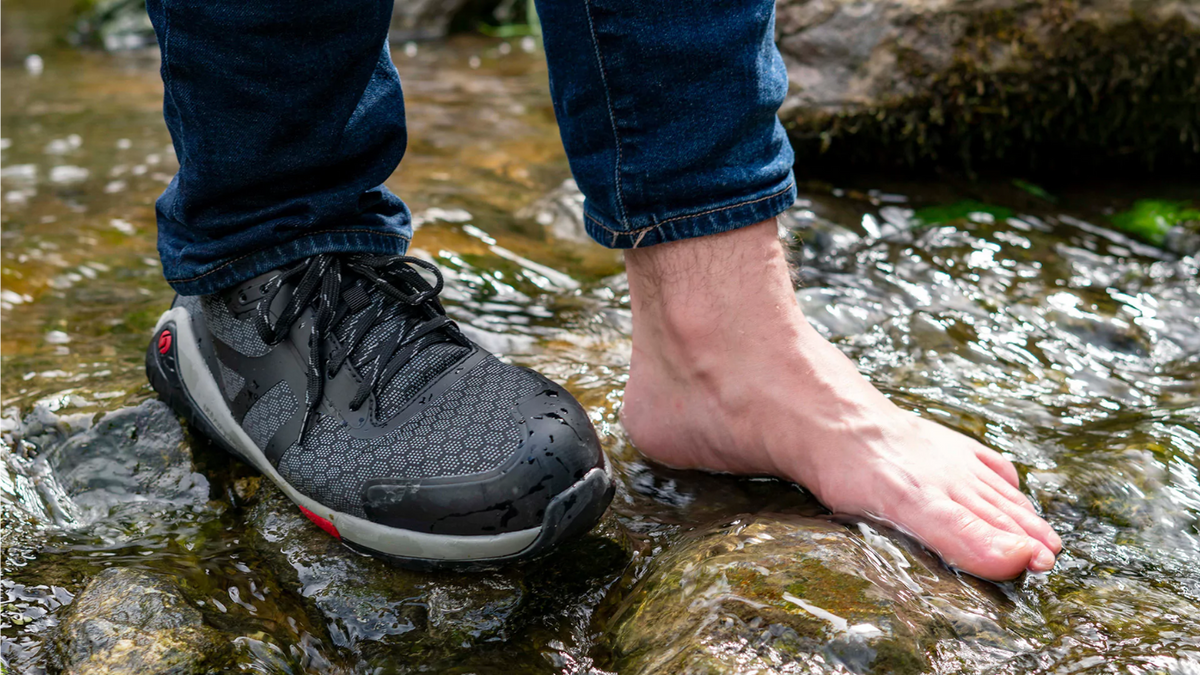 Barfuß-Sicherheitsschuh, Tragebild. Ein Fuß barfuß, ein Fuß im geschlossenen Schuh, Stand im flachen Gewässer.