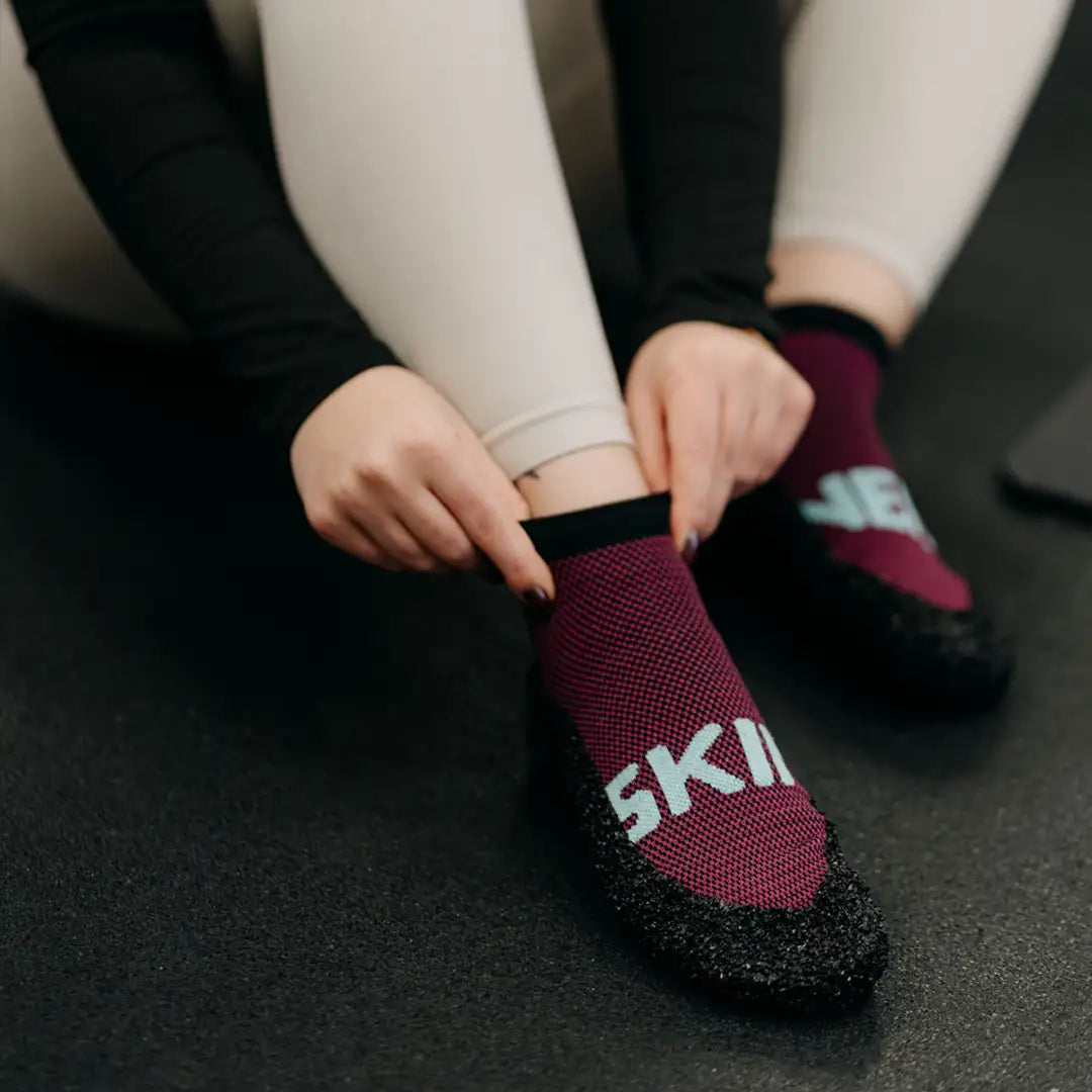 Person zieht bordeauxrote Skinners Socken an, Detailansicht von Material, Dehnbarkeit und Sitz am Fuß.
